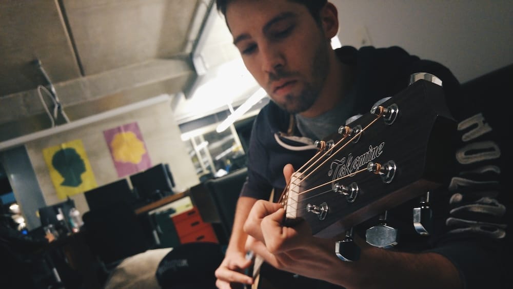 Homem jovem tocando violão Takamine em um ambiente interno, com foco nas mãos e na cabeça do violão, enquanto ele está concentrado na música.