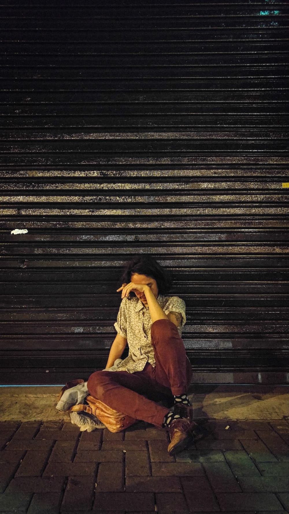 Pessoa sentada no chão em frente a uma porta metálica preta fechada, com a cabeça abaixada e o braço cobrindo o rosto, transmitindo uma sensação de cansaço ou tristeza. Ela está vestindo camisa clara, calça marrom e sapatos, com uma bolsa ao lado.