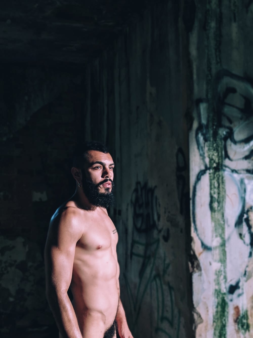 Homem pelado com barba e cabelo curto, olhando para o lado em um ambiente escuro com paredes grafitadas, iluminado por uma luz suave que destaca seu rosto e parte do corpo.