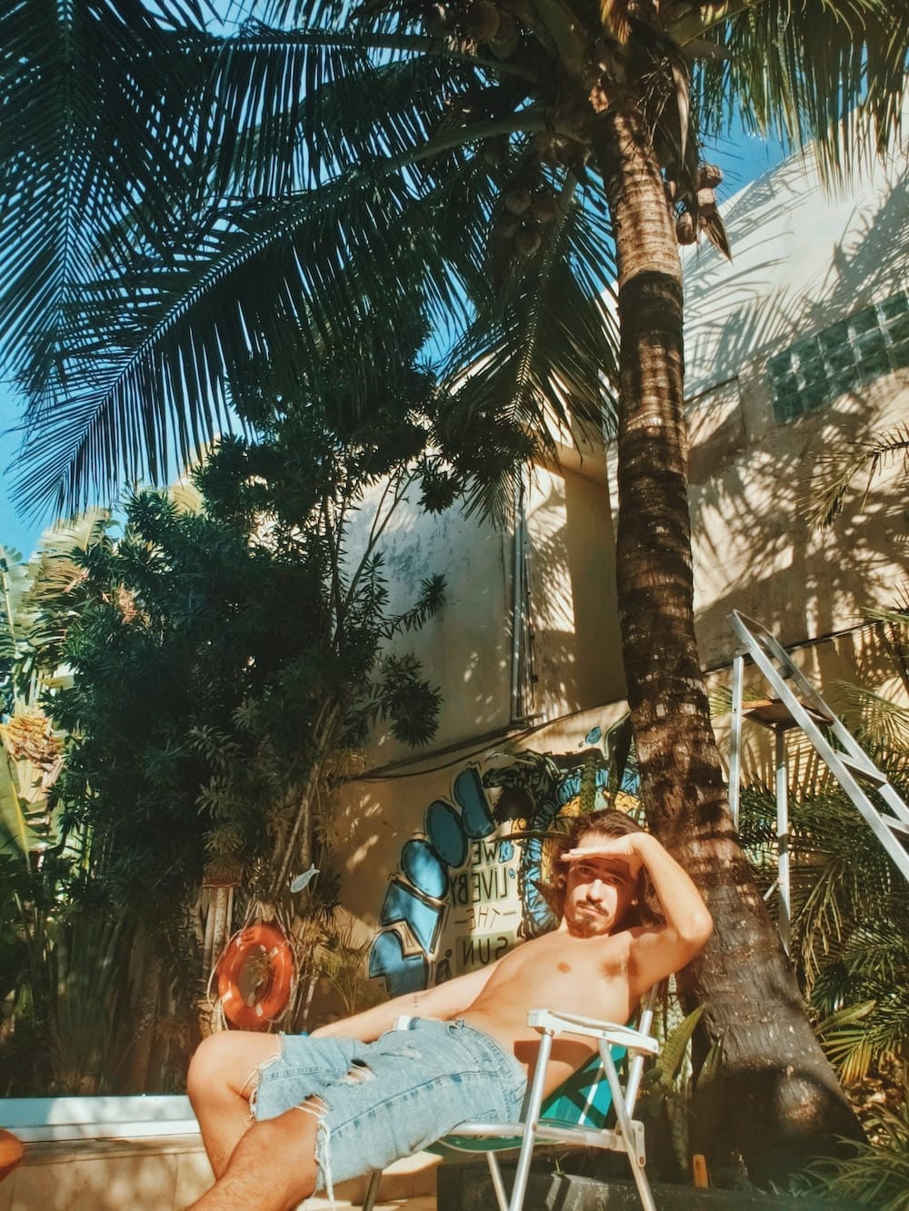 Homem sem camisa, usando shorts jeans rasgados, sentado em uma cadeira de praia sob uma palmeira, protegendo os olhos do sol com a mão, em um ambiente externo com vegetação e grafite na parede ao fundo.