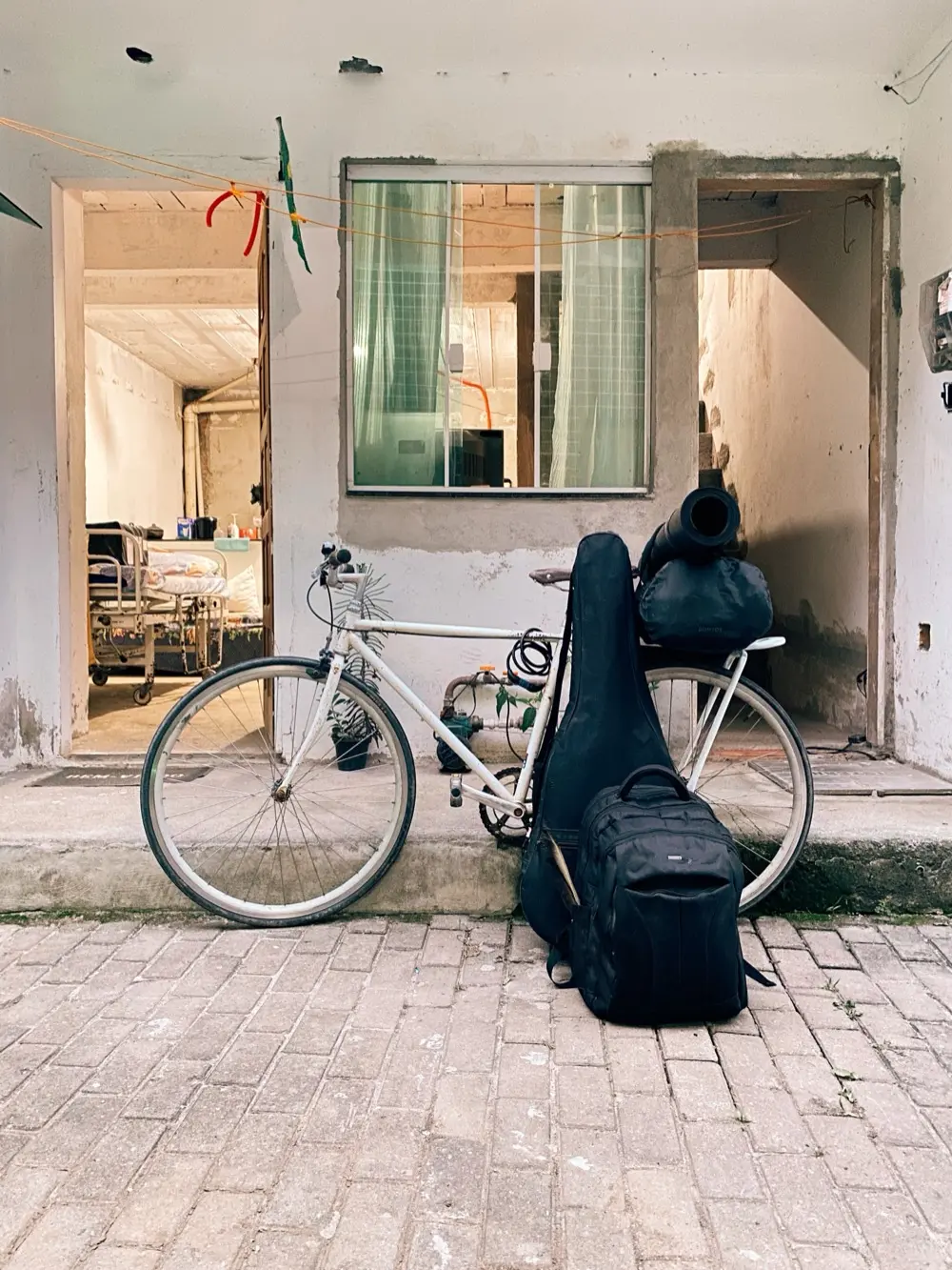 Bicicleta branca encostada em um pequeno degrau de uma construção inacabada; sobre o bagageiro há uma mala e um tapete de yoga e, apoiados na roda dianteira, um estojo de violão preto e uma mochila preta. Ao fundo, vê-se um quarto simples com uma cama hospitalar e paredes com reboco aparente. O chão é de paralelepípedos.