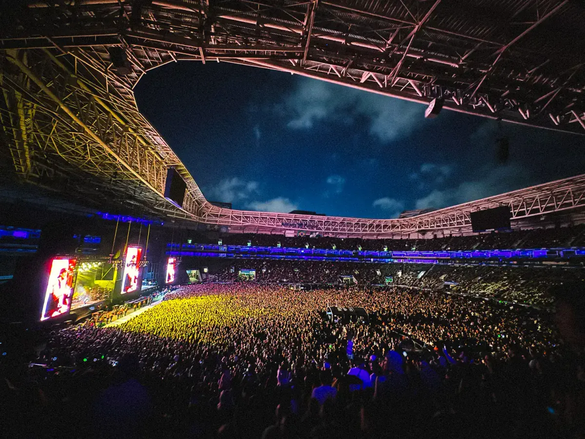 vista do Allianz durante festival I Wanna be Tour a noite com teto aberto revelando céu escuro e nuvens, multidão iluminada por luzes coloridas do palco, telões laterais exibindo performance