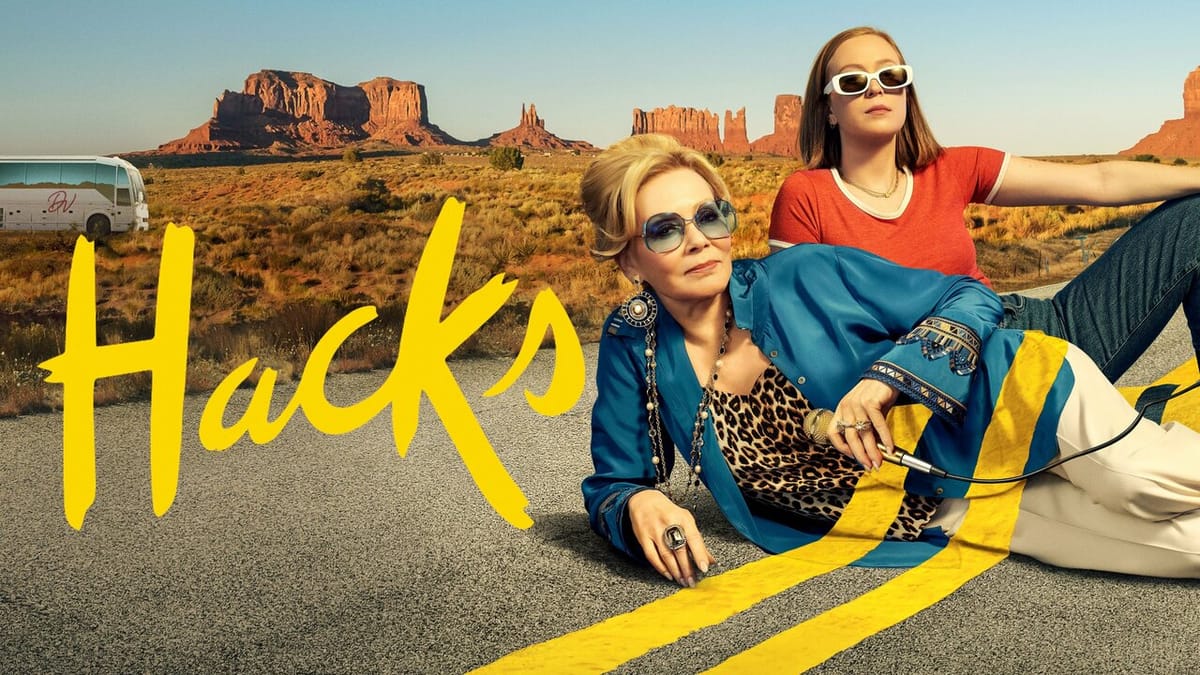 Pôster de "Hacks": duas mulheres deitadas em estrada no deserto com formações rochosas ao fundo. Uma loira de óculos escuros e jaqueta azul, outra jovem de blusa laranja.