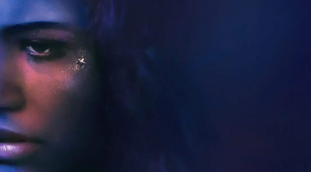 Rosto em close parcialmente iluminado por tons azuis e roxos. Maquiagem brilhante com silhueta de avião/pássaro na bochecha. Metade do rosto desaparece na escuridão.