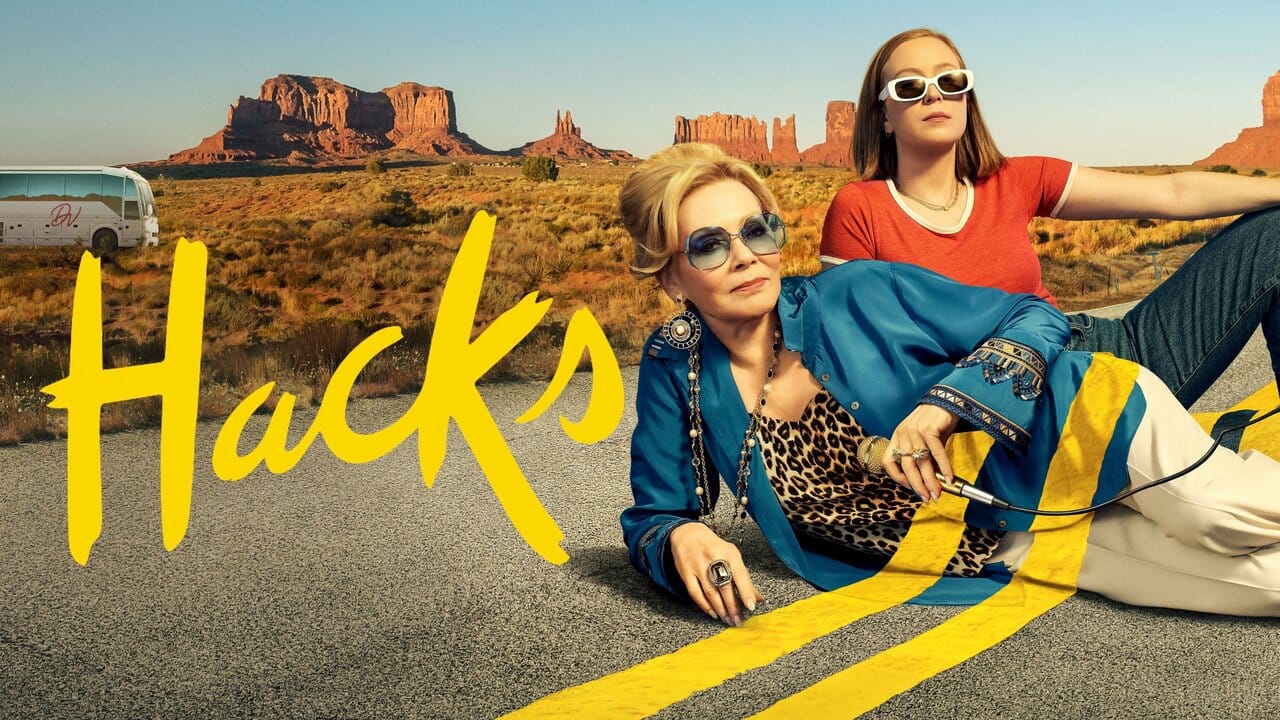 Pôster de "Hacks": duas mulheres deitadas em estrada no deserto com formações rochosas ao fundo. Uma loira de óculos escuros e jaqueta azul, outra jovem de blusa laranja.