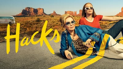 Pôster de "Hacks": duas mulheres deitadas em estrada no deserto com formações rochosas ao fundo. Uma loira de óculos escuros e jaqueta azul, outra jovem de blusa laranja.