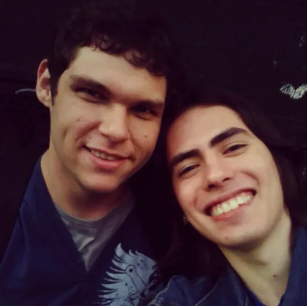 dois caras jovens tirando selfie, ambos sorrindo. o da esquerda tem cabelo cacheado escuro e usa uma camiseta, o da direita tem cabelo ondulado.